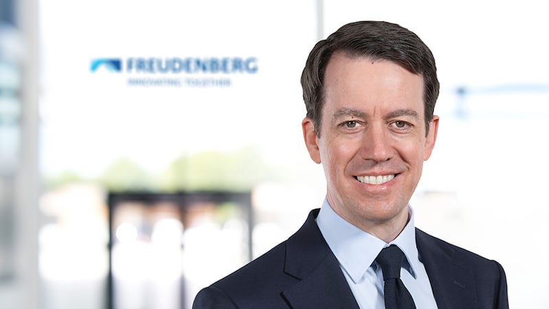 a headshot of a man in a suit and tie with a company logo blurred in the background